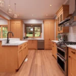 A modern cottage kitchen featuring shaker-style light wood cabinets, white subway tile backsplash, and white granite countertops. The space includes a large kitchen island with seating, a double oven range with a stainless steel hood, and warm hardwood flooring
